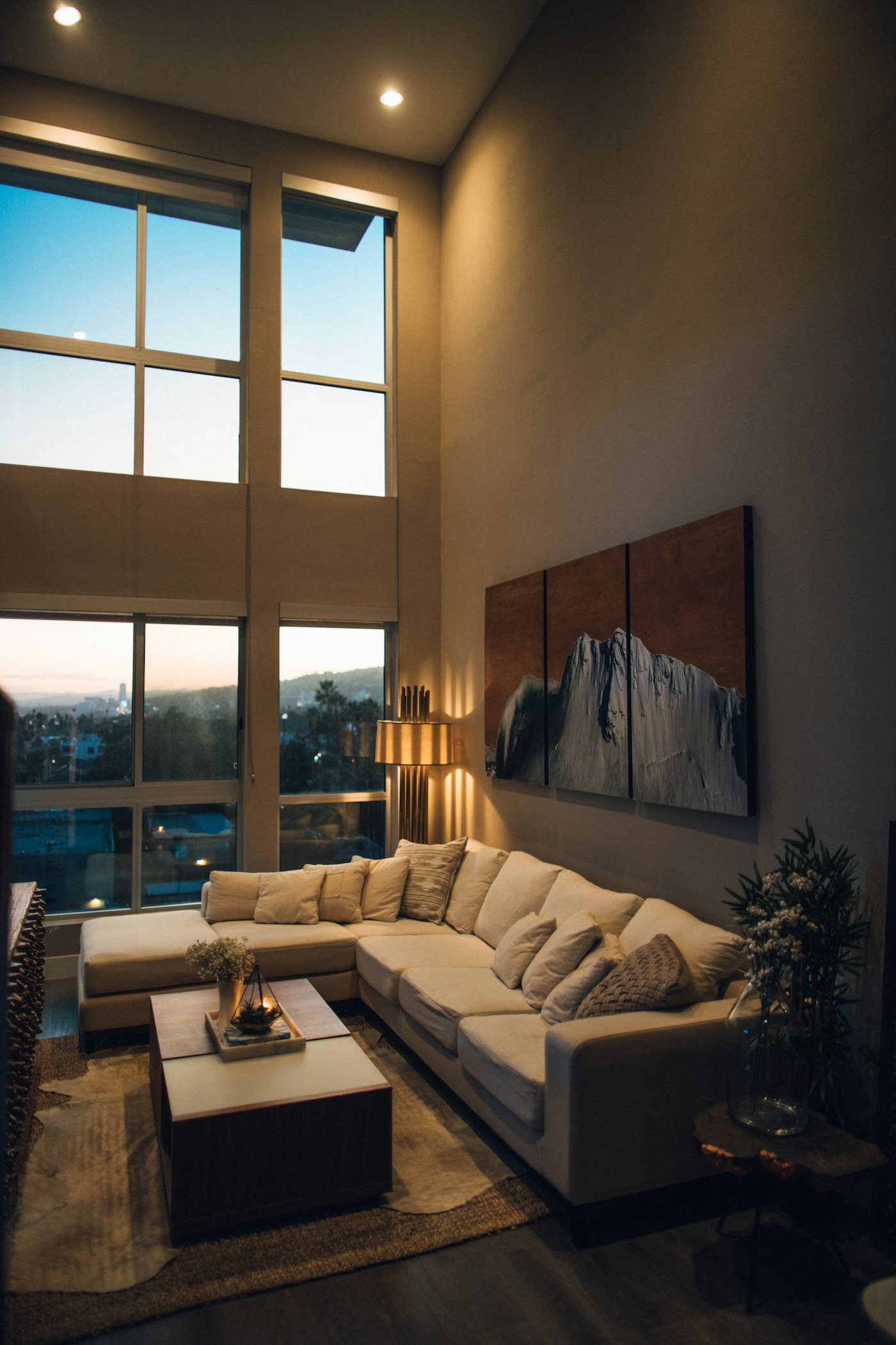 A double-height living room at dusk, a slim floor lamp and recessed downlights washing the wall above a pale corner sofa